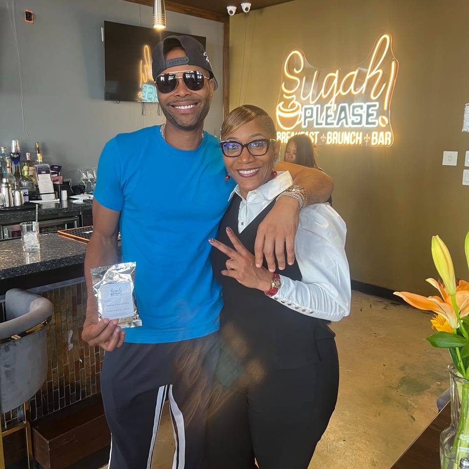 Two smiling people, a man and a woman, posing in front of a Sugah Please neon sign.