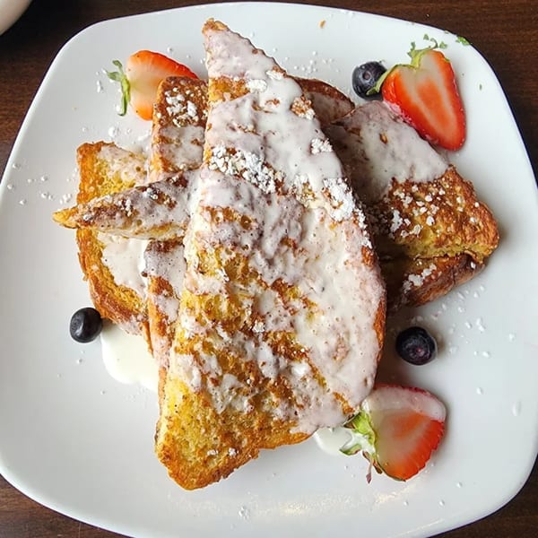 Sugah Please French toast with powdered sugar, glaze, strawberries, and blueberries on a white plate.