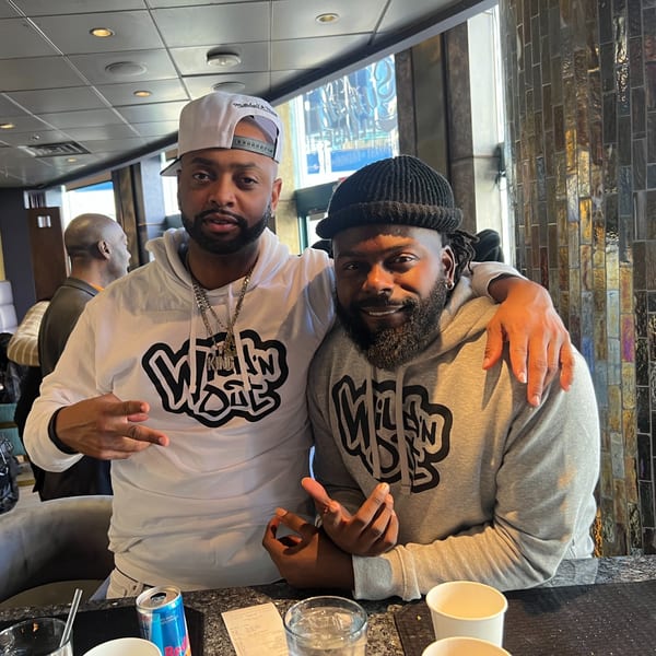 Two men in hoodies and hats pose at a bar, one with an arm around the other.