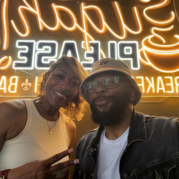 Two people smiling for a selfie in front of a bright neon sign at Sugah Please.