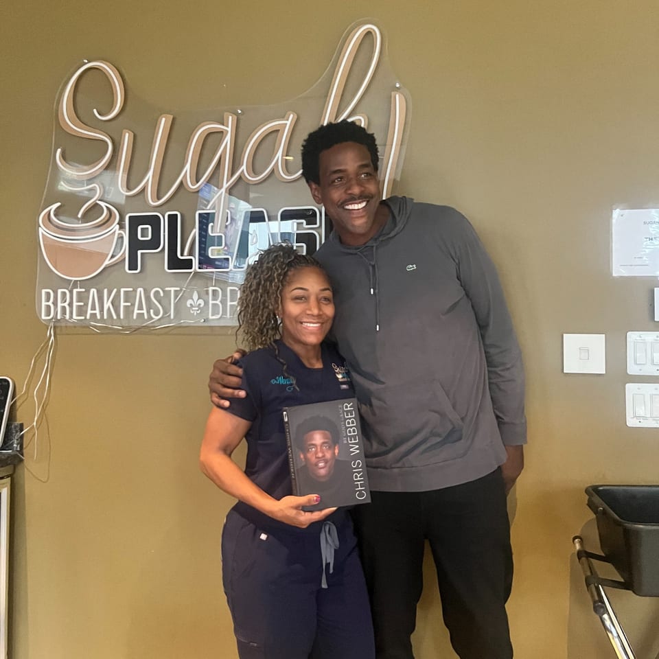 A woman and a man smiling, posing with a book in front of the "Sugah Please" sign.