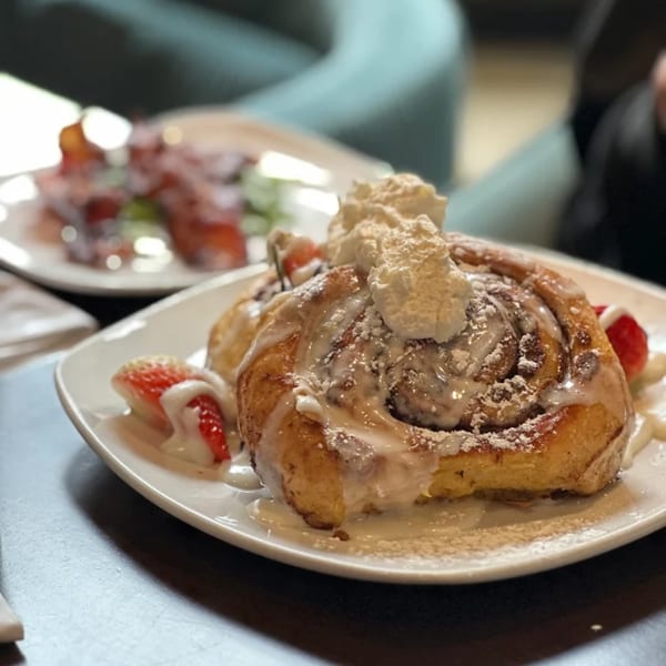 A large cinnamon roll with white icing, whipped cream, powdered sugar, and strawberries on a white plate.