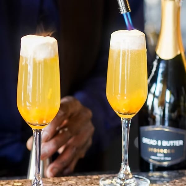 Two Sugah Please mimosas with foamy tops, one being torched, and a bottle of sparkling wine.