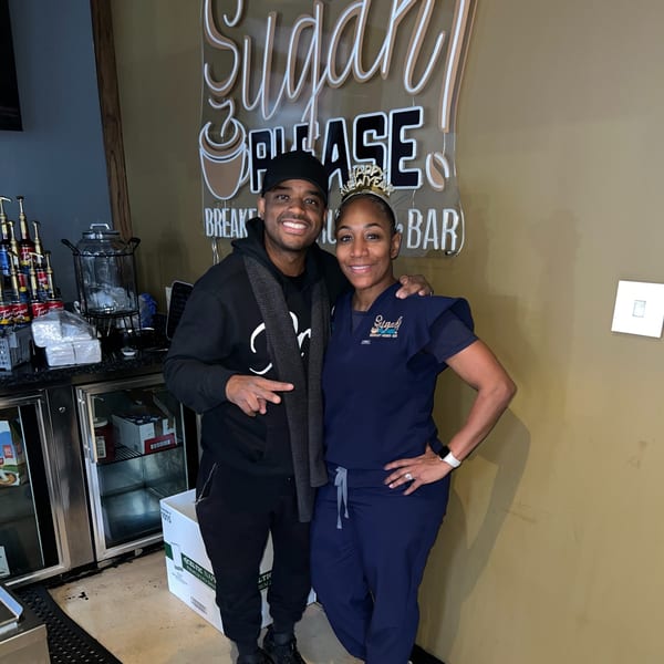 Two people smiling in front of a neon "Sugah Please Breakfast Brunch Bar" sign.