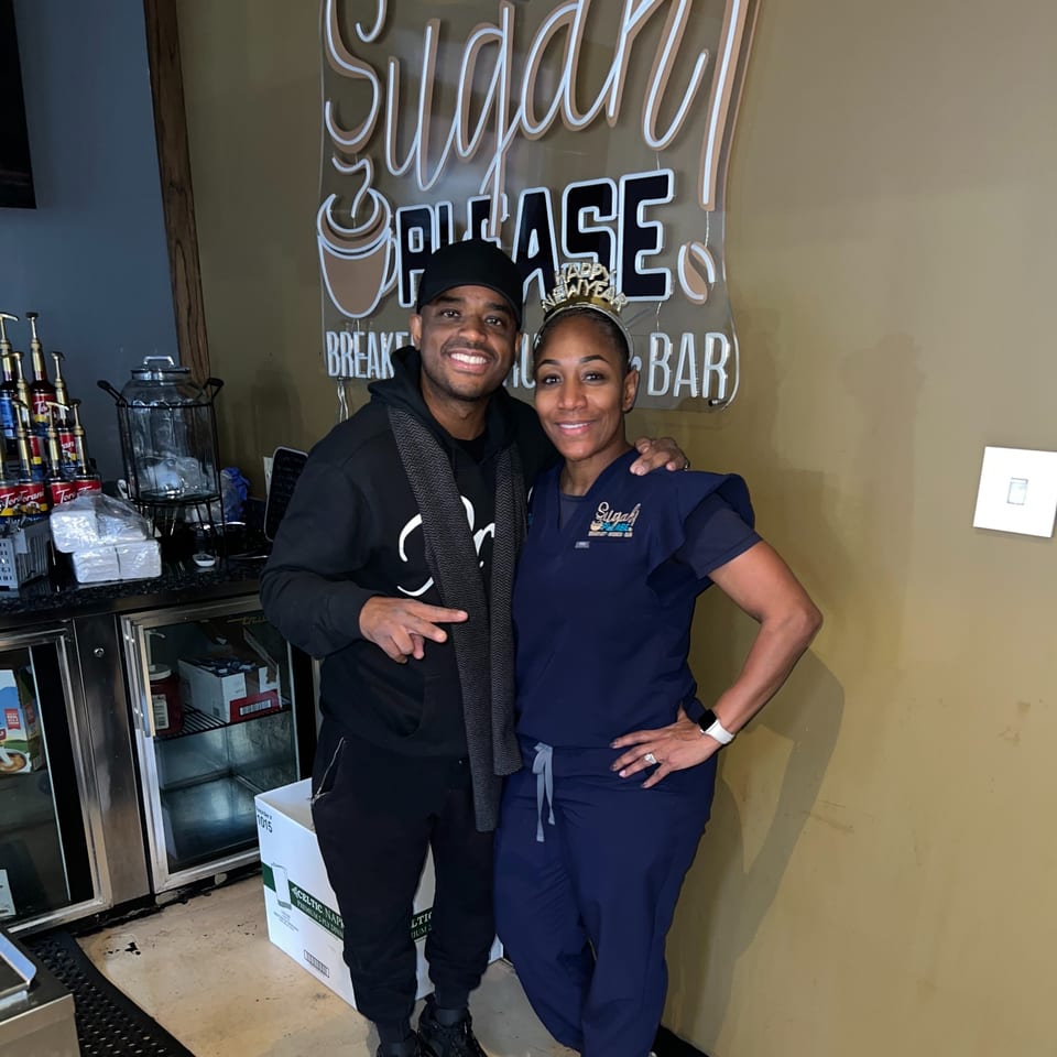 Two people smiling in front of a neon "Sugah Please Breakfast Brunch Bar" sign.