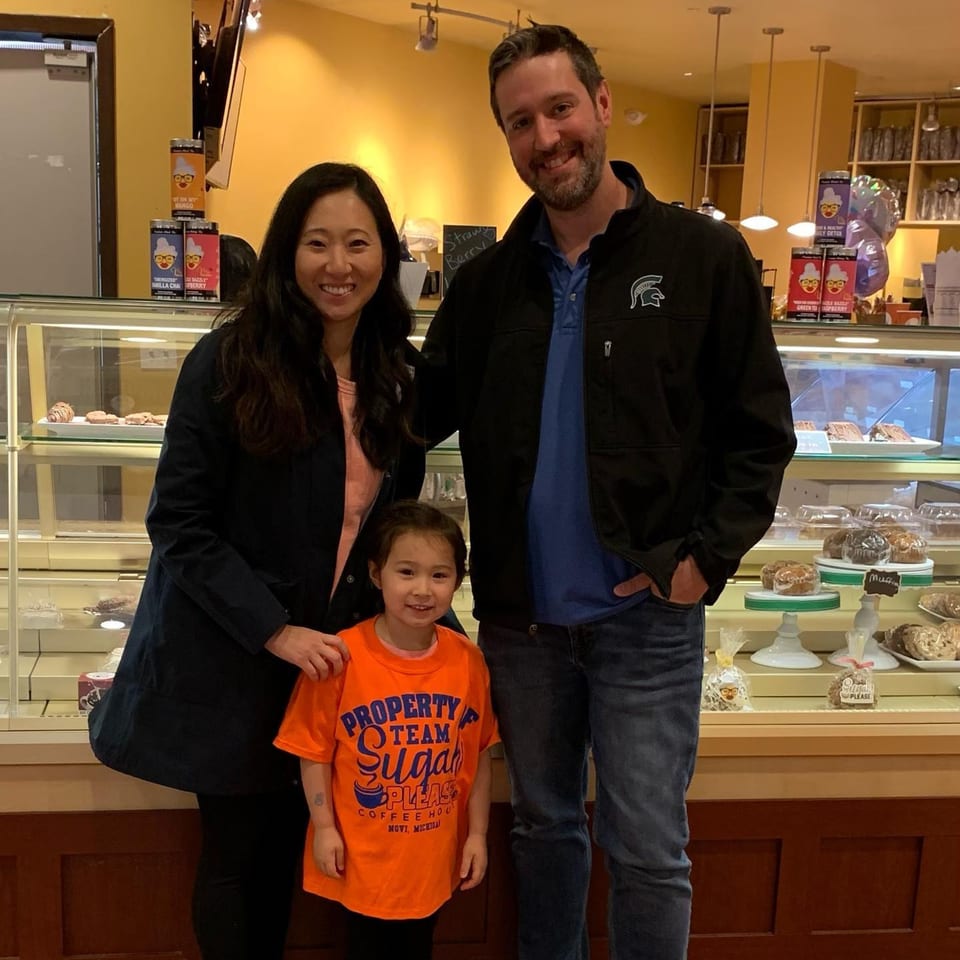 A family of three, including a young girl in a "Sugah Please" t-shirt, smiling in front of a pastry display.