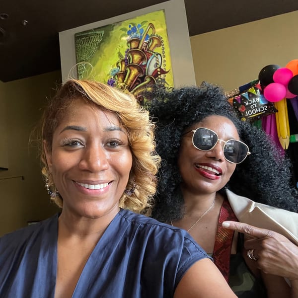 Two women smiling at Sugah Please, one pointing, with a colorful musical instrument painting in the background.