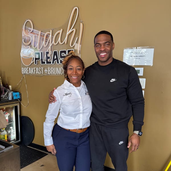 Two smiling people, a woman and a man, stand in front of the "Sugah Please Breakfast Brunch" sign.
