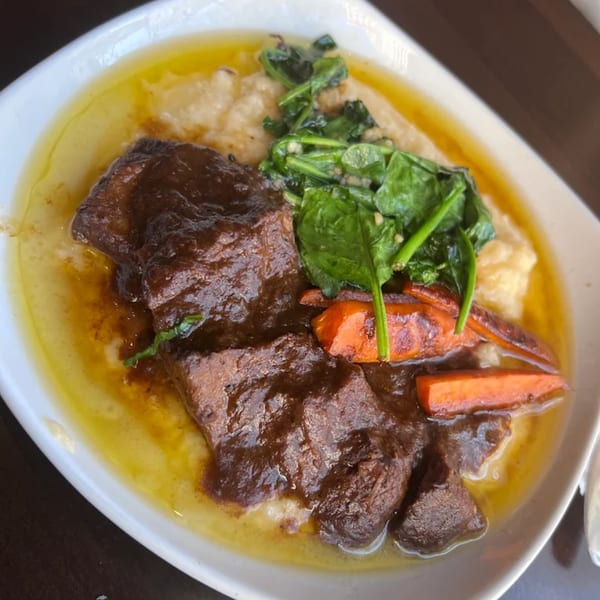 Braised short ribs with creamy grits, sautéed spinach, and roasted carrots in a white bowl.