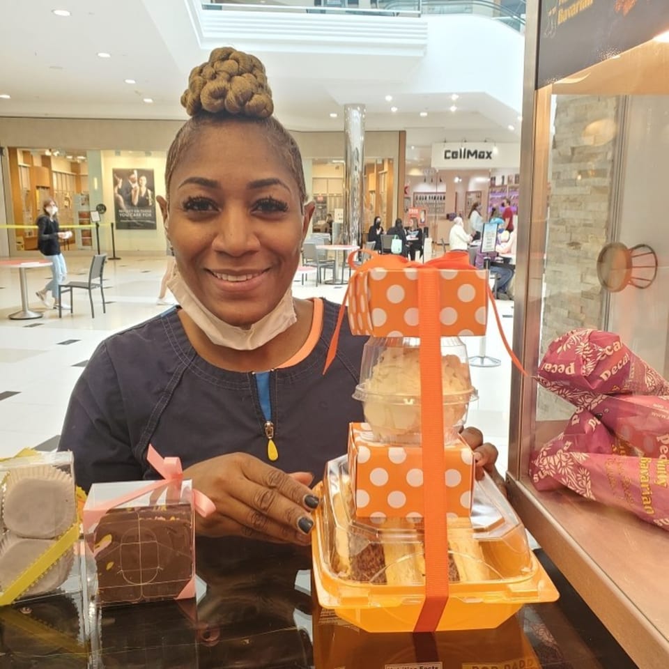 Smiling woman with braided hair bun behind a counter with Sugah Please desserts in a mall.