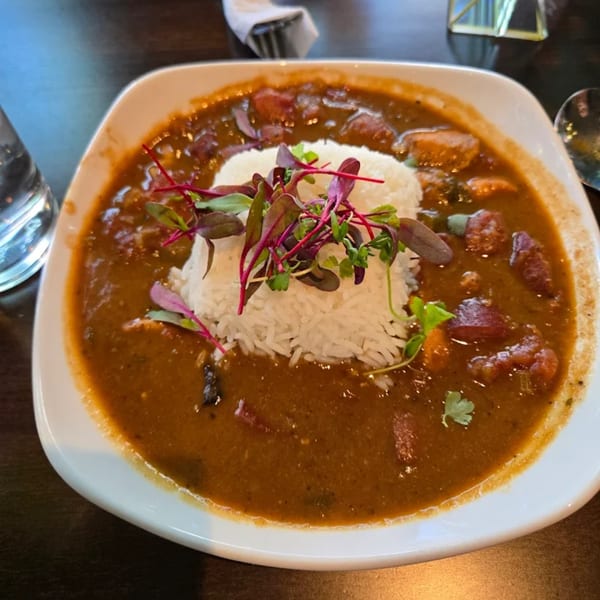 A white bowl of rich brown gumbo with rice and microgreens, served at Sugah Please.
