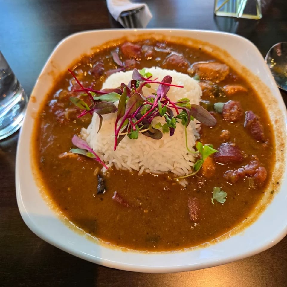 A white bowl of rich brown gumbo with rice and microgreens, served at Sugah Please.