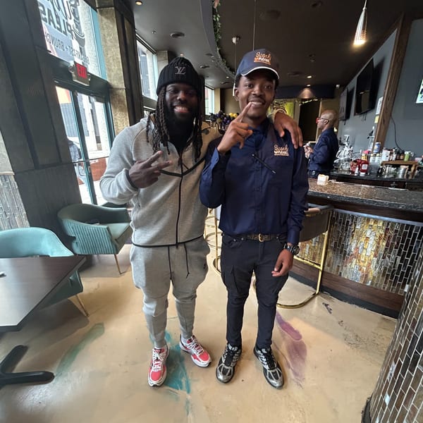 Two men smiling and posing inside Sugah Please Breakfast Brunch Bar, one wearing a branded cap.